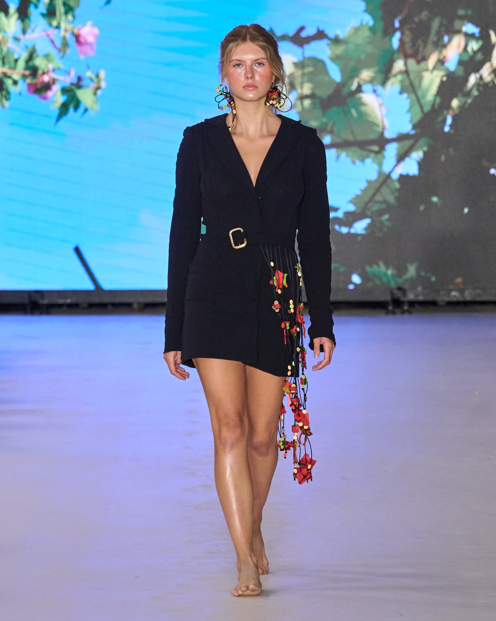 A model walking the runway wearing a black blazer and black skirt with floral detailing.