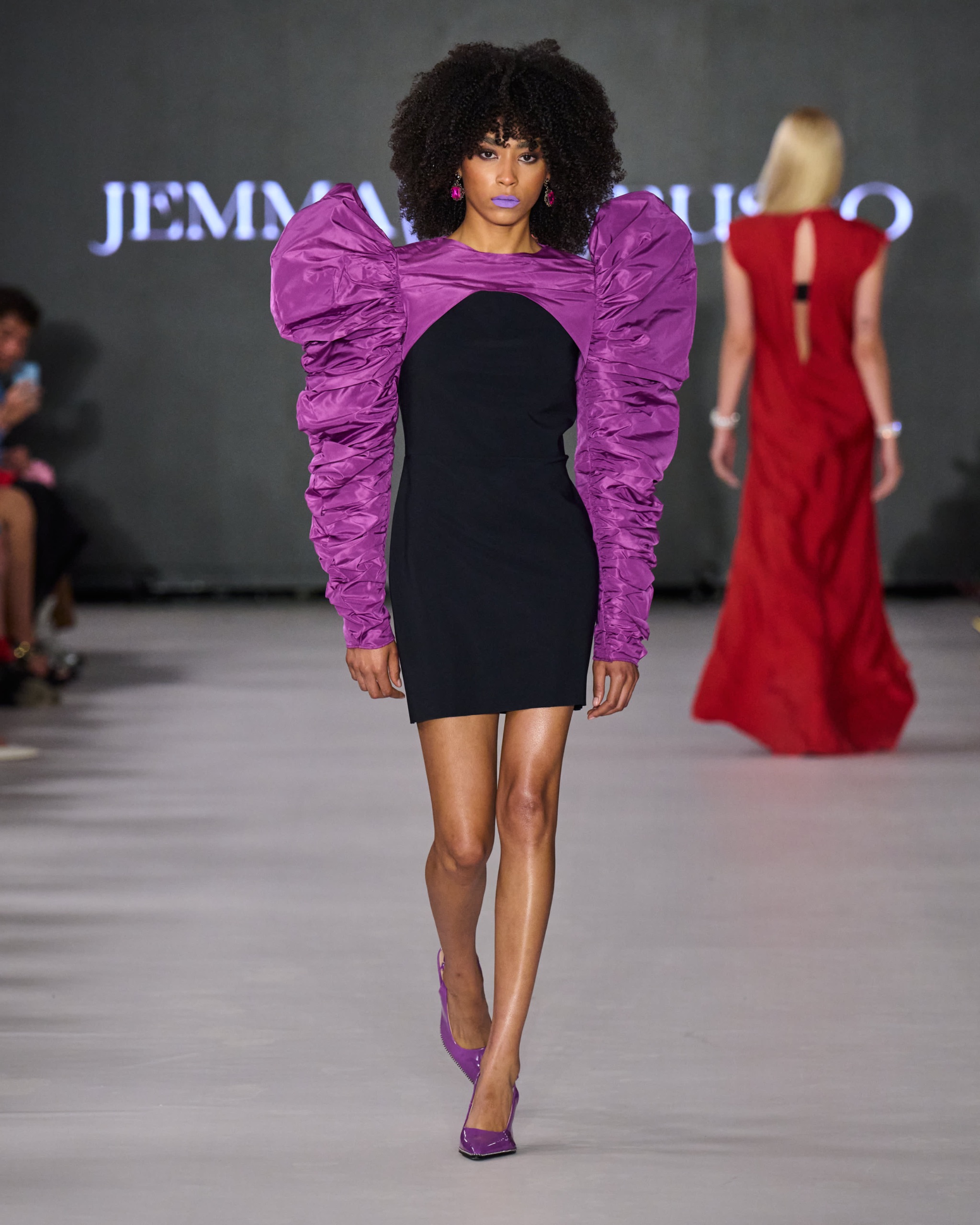 A model walking the runway wearing a short black dress with purple sleeves.